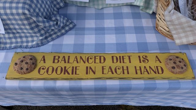 A yellow wooden sign on a blue gingham market table that reads: A balanced diet is a cookie in each hand.